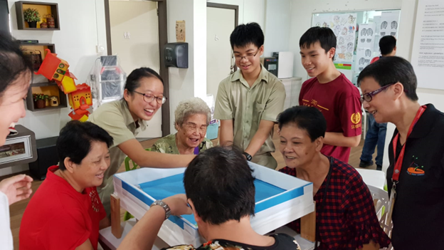 A group of people of mixed ages gather around a white tray with blue water, smiling indoors.